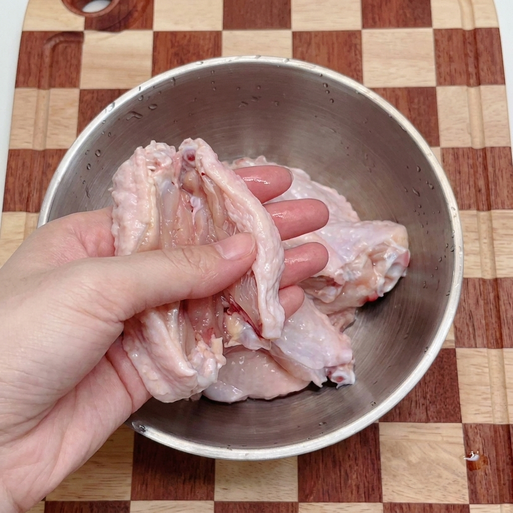 Unas manos usando tijeras de cocina para cortar alitas de pollo crudas en dos piezas dentro de un bol de acero inoxidable.