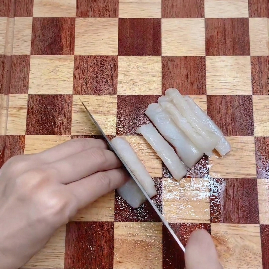 Un cuchillo de cocina cortando un filete de bacalao blanco en tiras uniformes sobre una tabla de cortar de madera.
