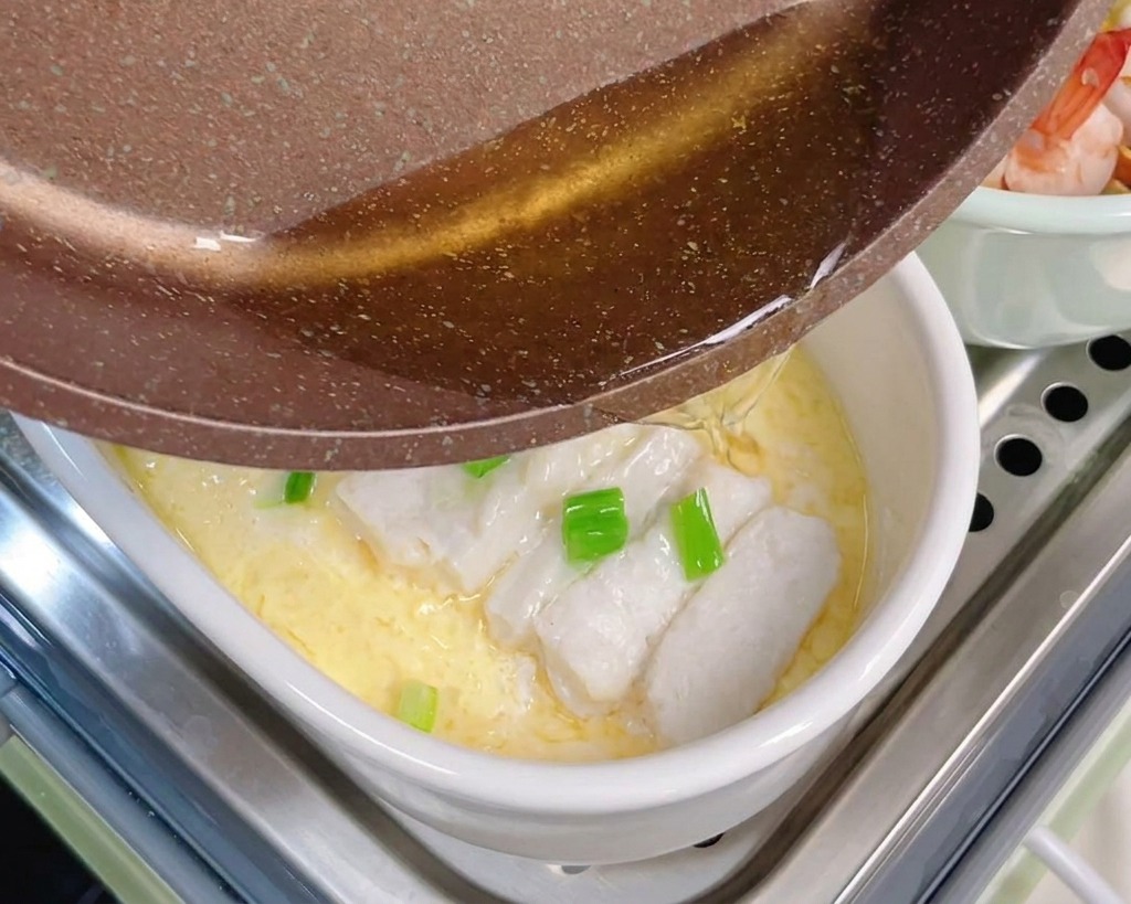 Aceite caliente vertiéndose desde una sartén marrón sobre cebolletas picadas y bacalao al vapor encima de un huevo suave.