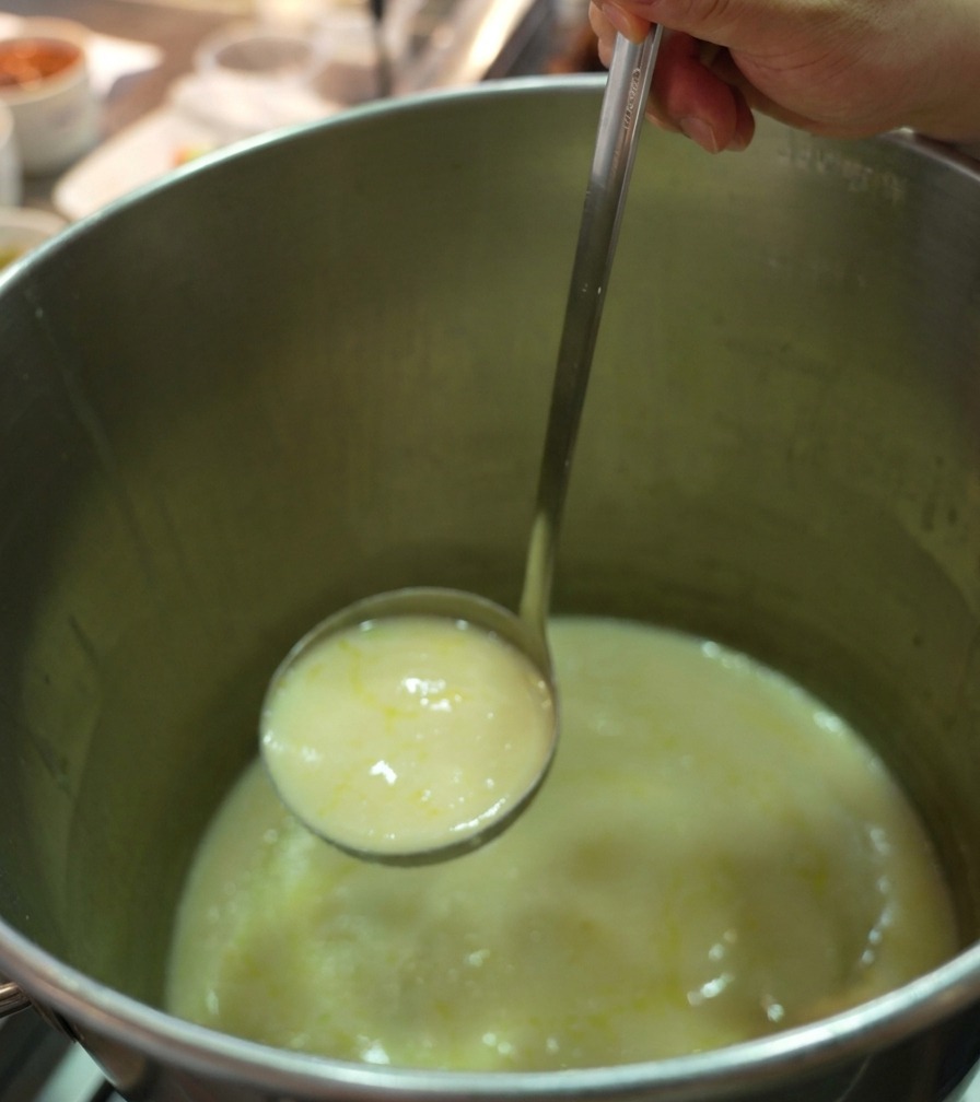 Un cucharón de acero inoxidable levantando un caldo de pollo espeso y cremoso de una olla profesional.