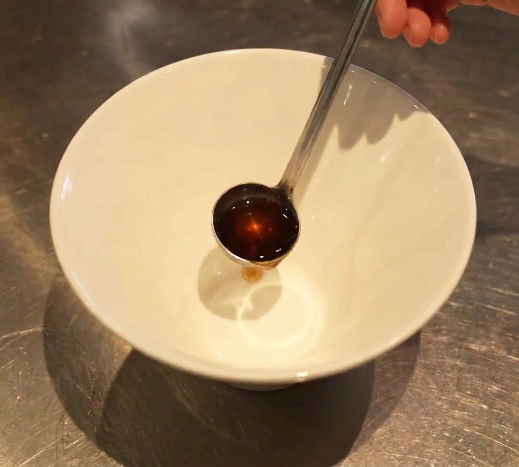 Un pequeño cucharón vertiendo un sazonador líquido oscuro en el fondo de un bol de ramen blanco vacío.