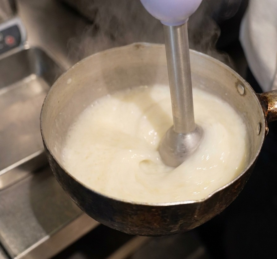 Una batidora de mano sumergida en un cazo con líquido amarillo, creando una espuma blanca y espesa en la superficie.