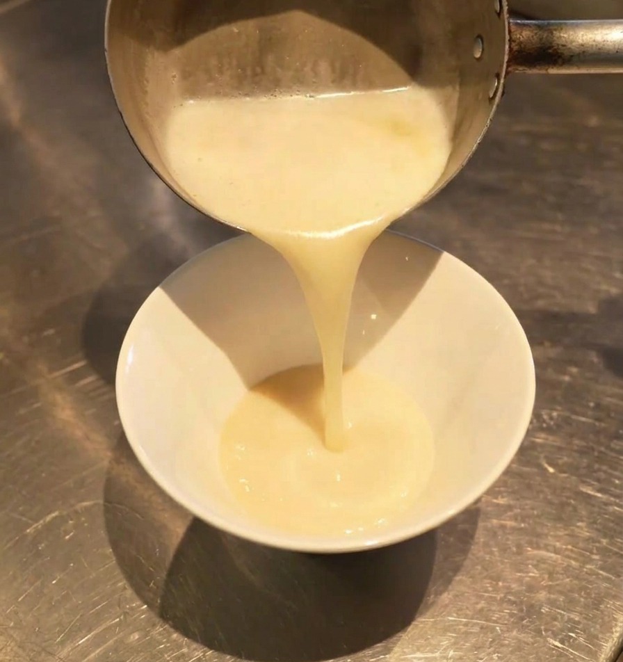 Caldo blanco lechoso vertiéndose desde un cazo en un bol blanco, mezclándose con el sazonador oscuro del fondo.