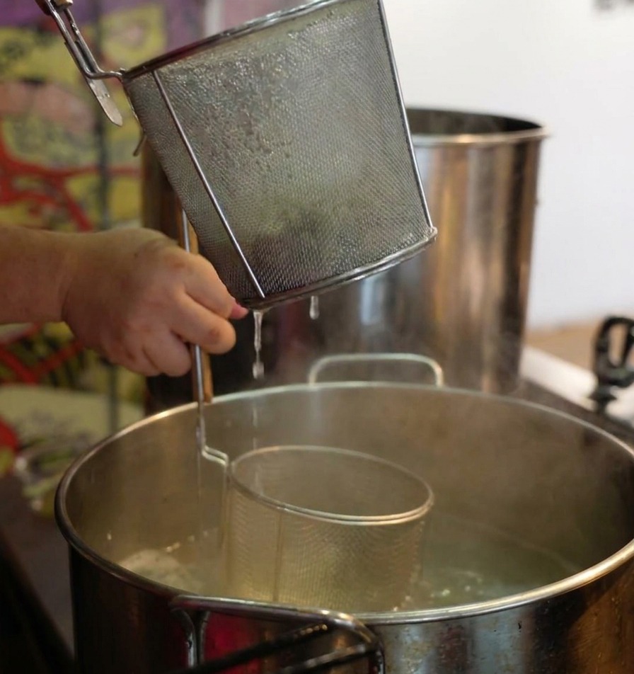 Manos sujetando un colador de fideos sobre una olla humeante, agitándolo para escurrir los fideos ramen cocidos.