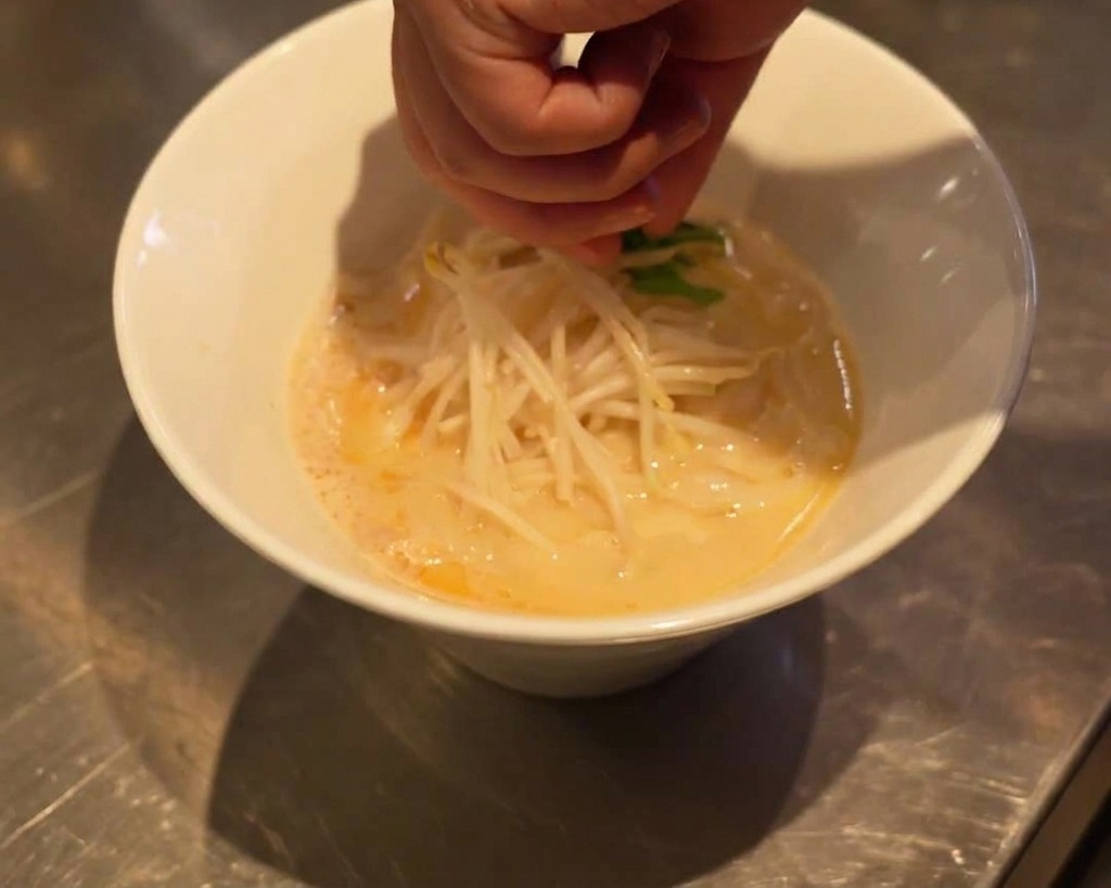 Una mano colocando cuidadosamente hojas de espinaca fresca sobre los brotes de soja en el bol de ramen.