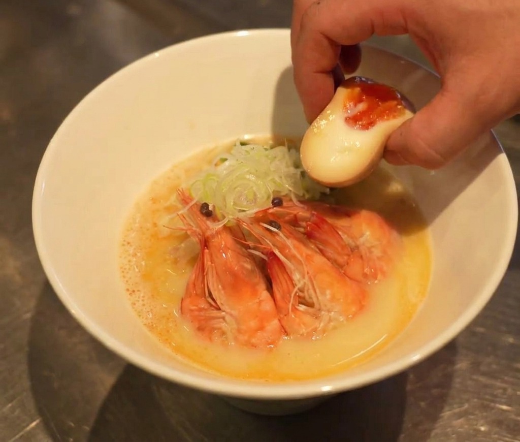 Una mano colocando medio huevo pasado por agua con la yema líquida en un bol de ramen casi terminado.