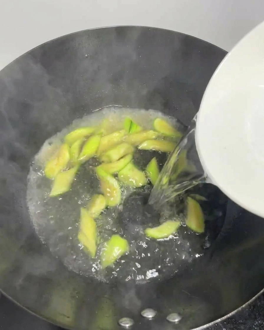 Agua clara vertiéndose desde un tazón blanco en un wok burbujeante con rodajas de lufa.
