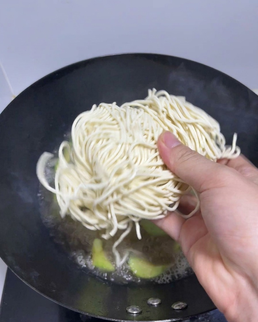 Una mano colocando un nido de fideos ramen frescos en un wok con caldo hirviendo.