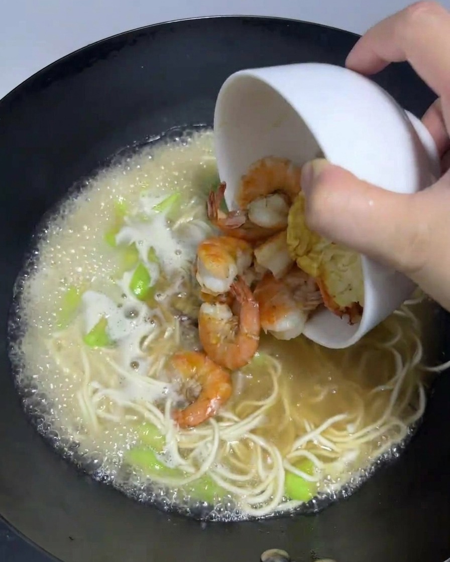 Camarones cocidos y un huevo frito vertiéndose de nuevo en el wok con el ramen hirviendo.