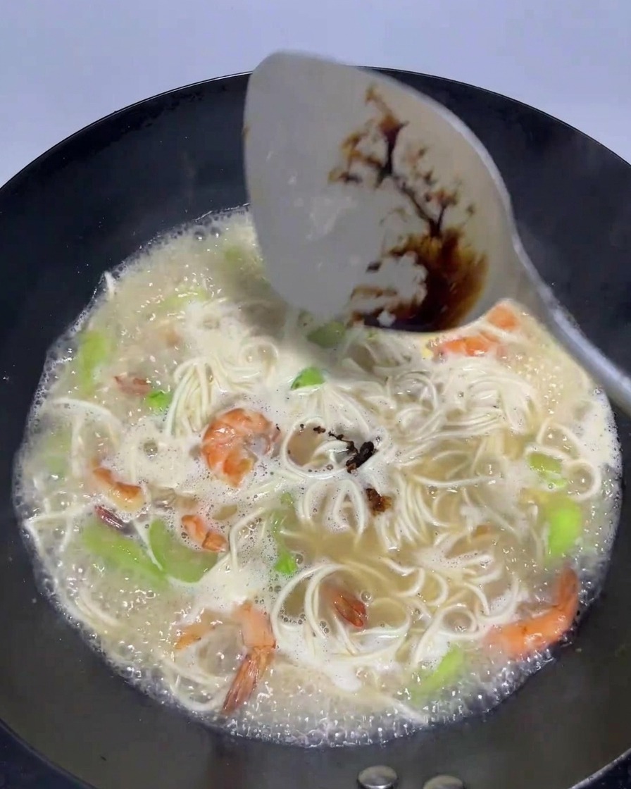Una espátula vertiendo salsa de soja oscura en un wok con caldo burbujeante, fideos y mariscos.