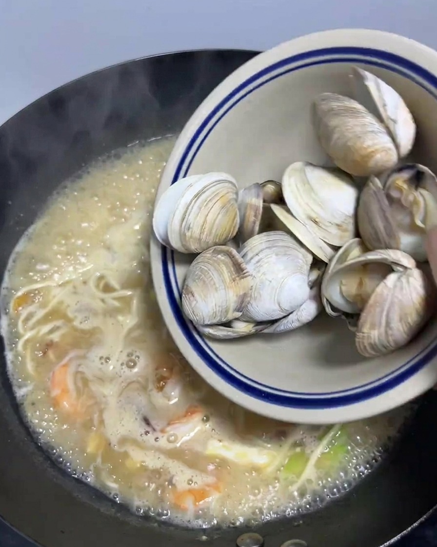 Un tazón de almejas blanqueadas vertiéndose en un wok grande lleno de ramen de mariscos.