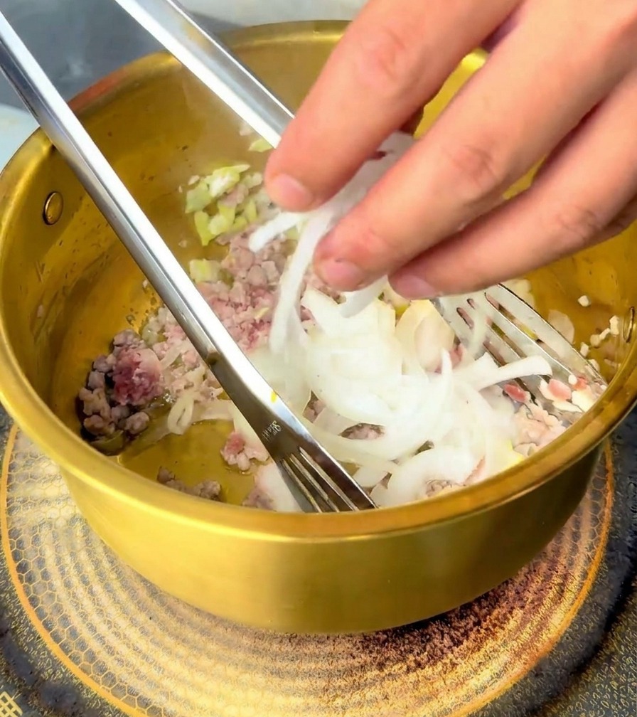 Una mano añadiendo rodajas de cebolla blanca a la carne de cerdo salteada en la olla.