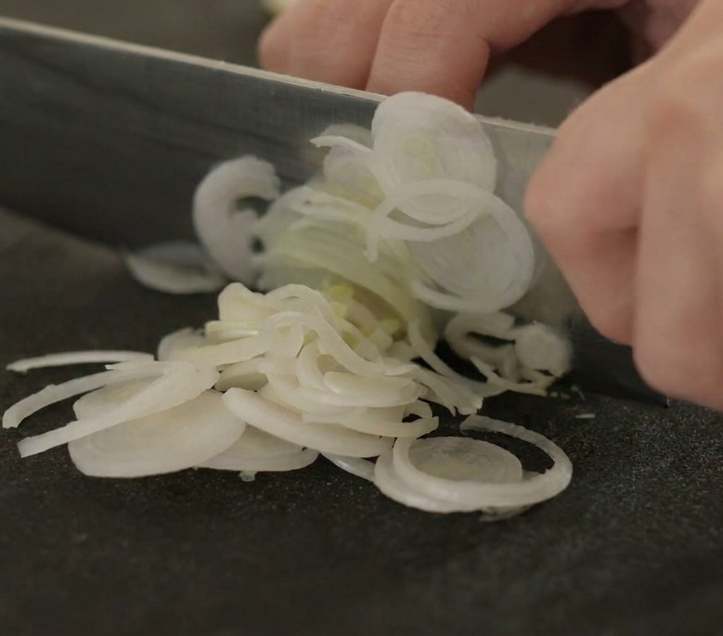 Primer plano de unas manos usando un cuchillo de chef para cortar puerros en rodajas finas sobre una tabla negra.