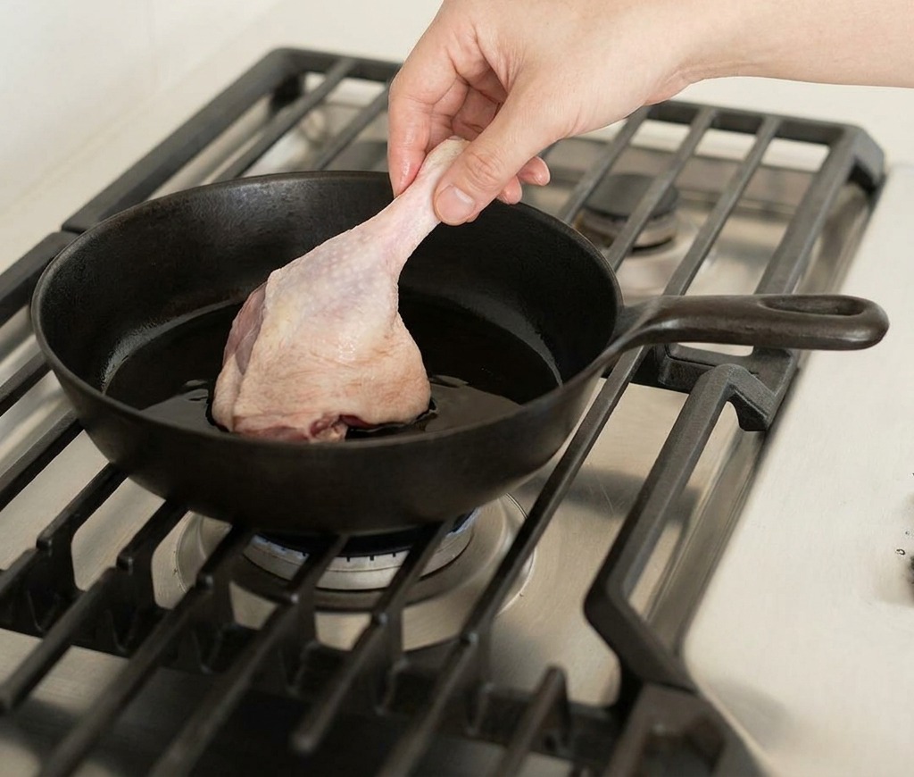 Una mano colocando un muslo de pato crudo en una sartén de hierro fundido caliente sobre un quemador de gas.
