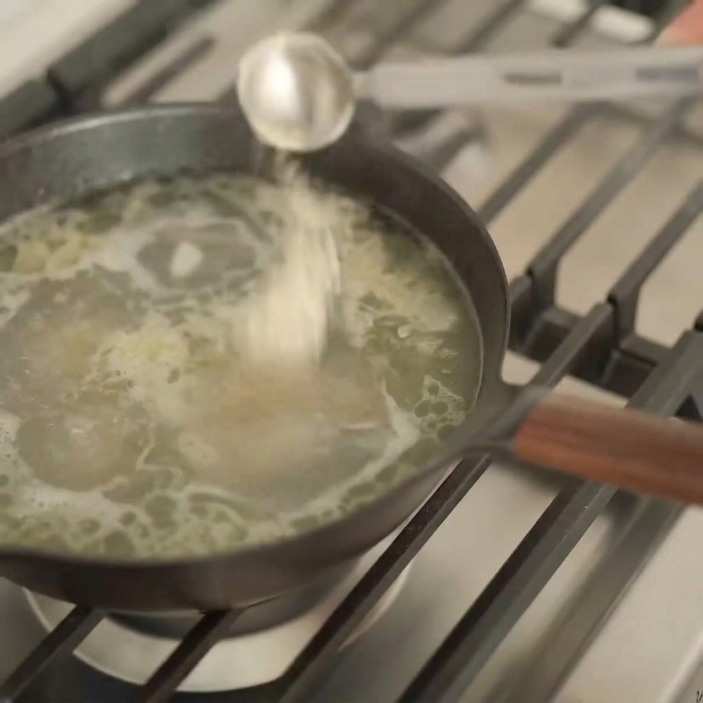 Una cuchara medidora de plata vertiendo un líquido oscuro en una olla con caldo de ramen de color claro.