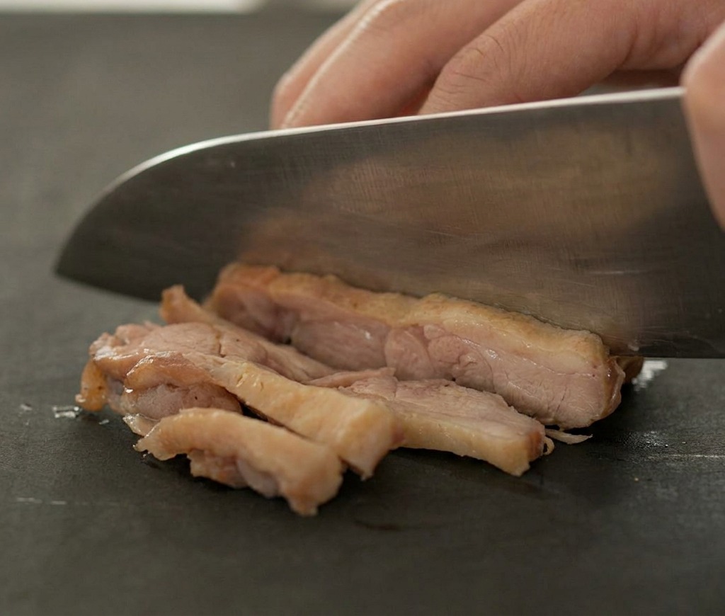 Un cuchillo afilado cortando una pieza de carne de pato dorada sobre una tabla de cortar oscura.