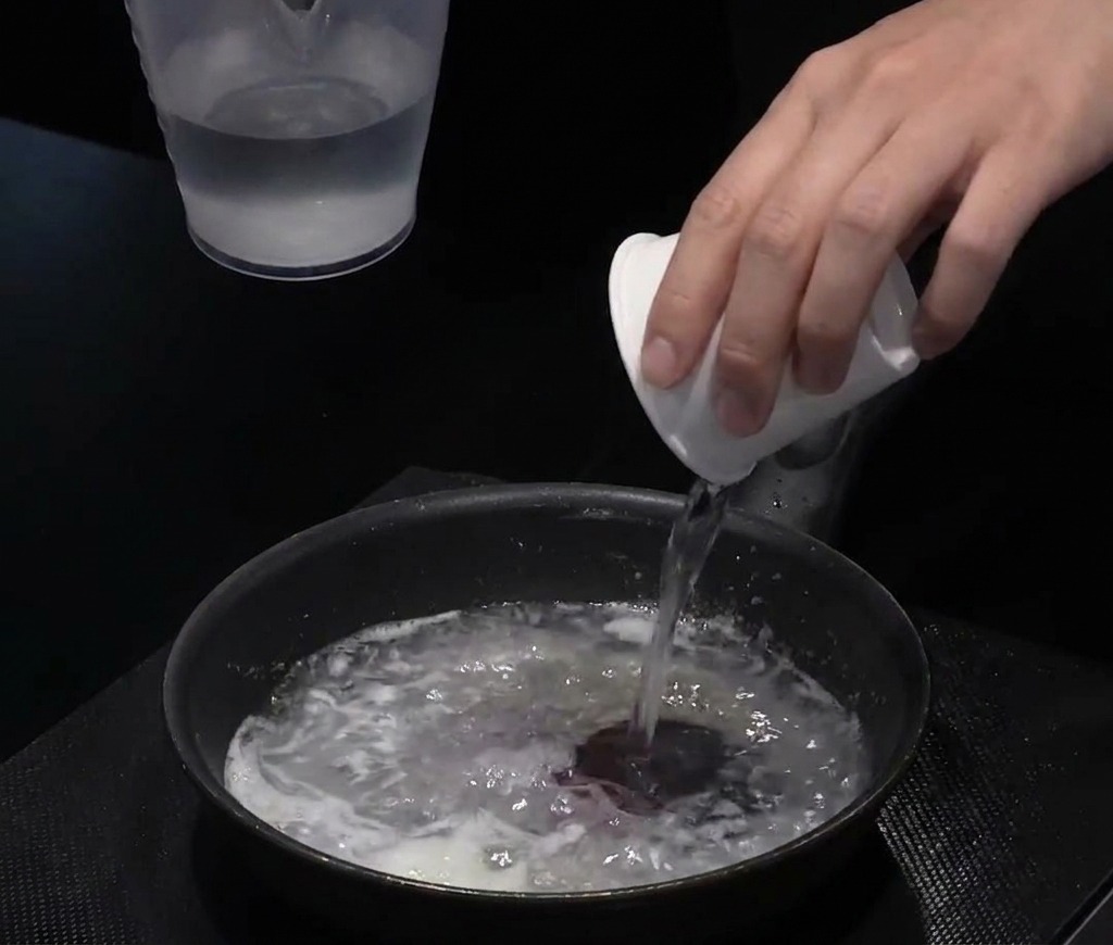 Vertiendo agua desde una taza blanca en una sartén negra con la mezcla de aceite y harina.