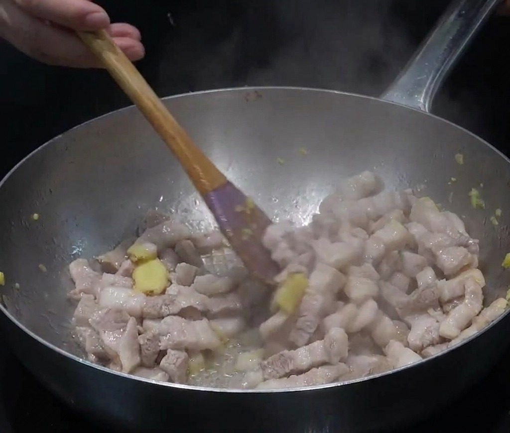 Rodajas de panceta de cerdo y jengibre picado salteándose en una olla de acero inoxidable con vapor.