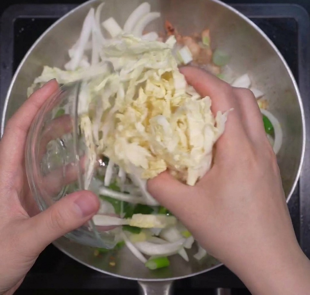 Manos añadiendo una gran porción de col napa rallada a una olla con cebollas y cebolletas salteadas.