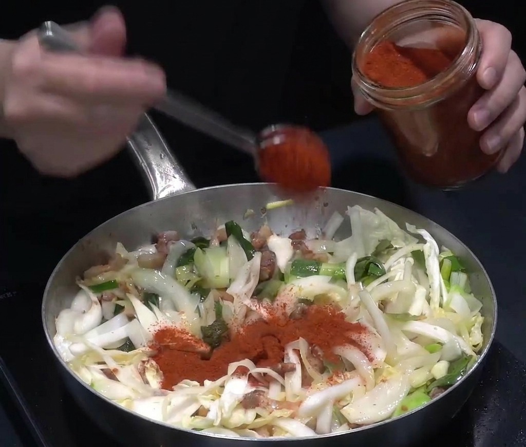 Polvo de chile rojo intenso cayendo desde una cuchara sobre la mezcla de carne y verduras en la olla.