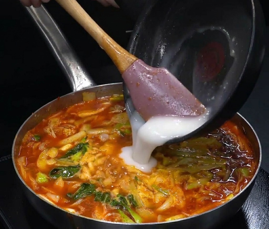 Un líquido opaco blanquecino vertiéndose desde una sartén pequeña a una olla grande de sopa roja burbujeante.