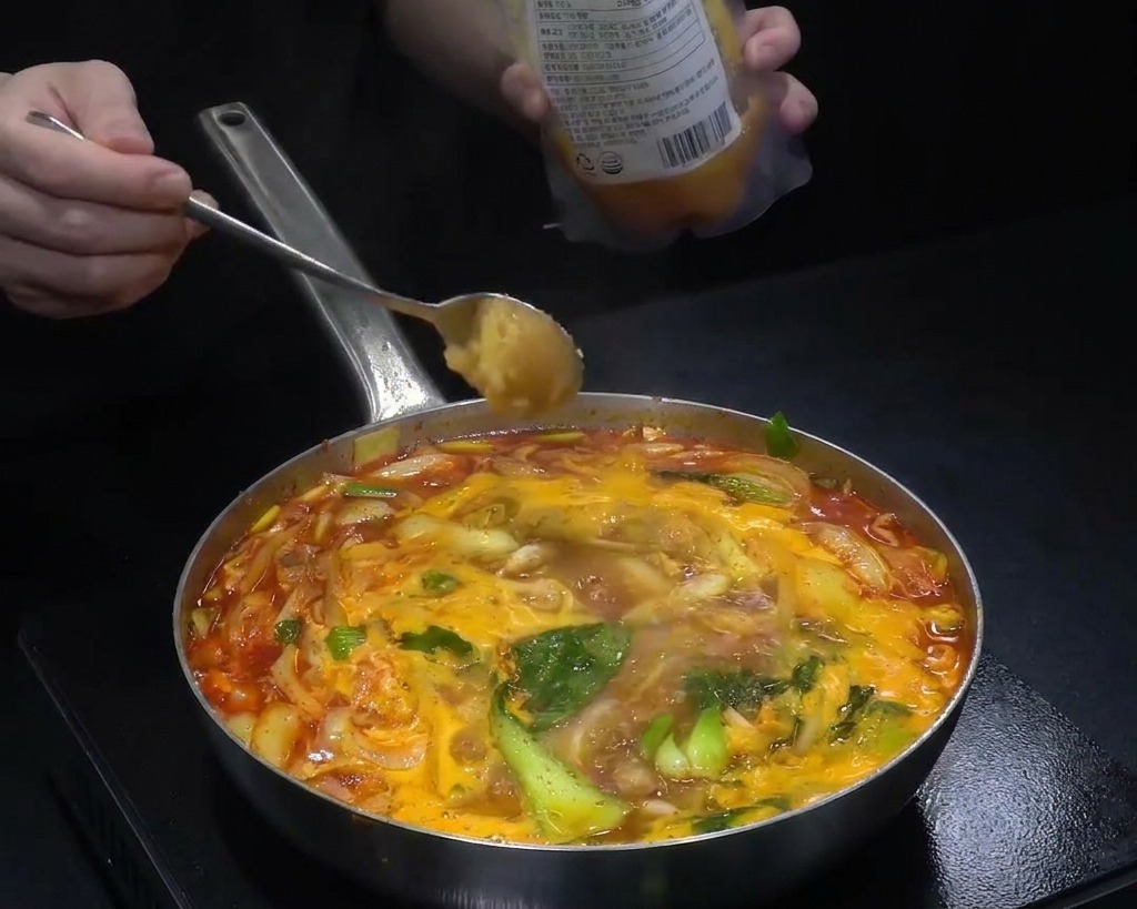 Una cucharada de ajo picado añadiéndose a una olla de sopa roja hirviendo llena de col y cebolla.