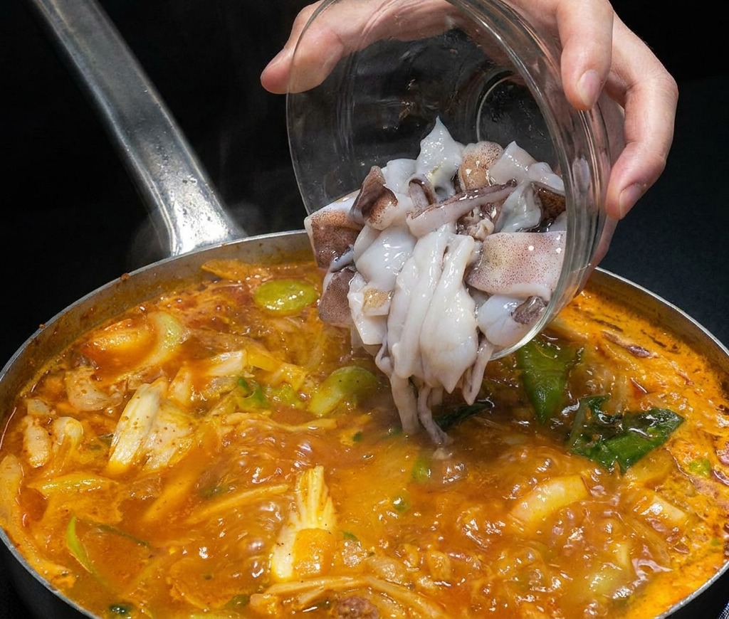 Trozos de calamar crudo vertiéndose desde un bol de cristal a una olla de caldo de mariscos humeante.