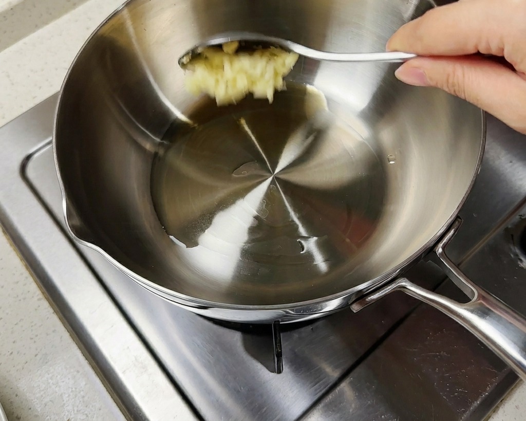 Una cuchara con ajo picado añadiéndose a una olla que contiene aceite caliente y cebolletas verdes picadas.