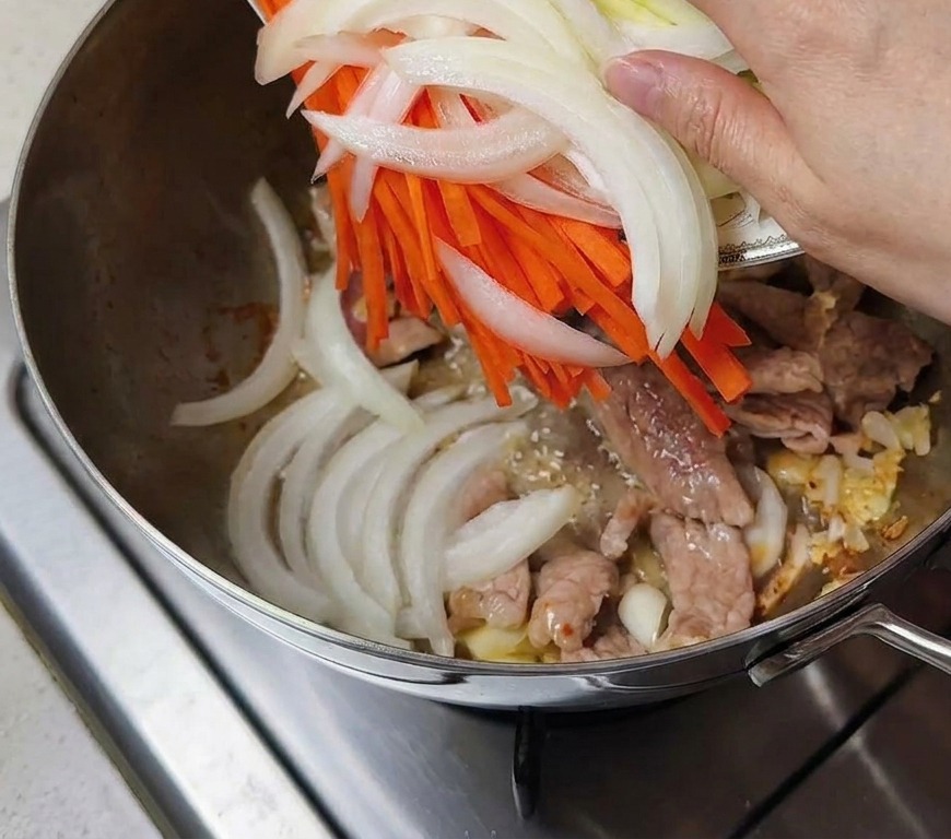 Mano vertiendo un plato de cebolla, zanahoria y calabacín en la olla con la carne.