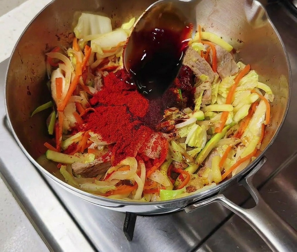 Cuchara de plata vertiendo salsa de soja oscura sobre verduras con chile en una olla de acero inoxidable.