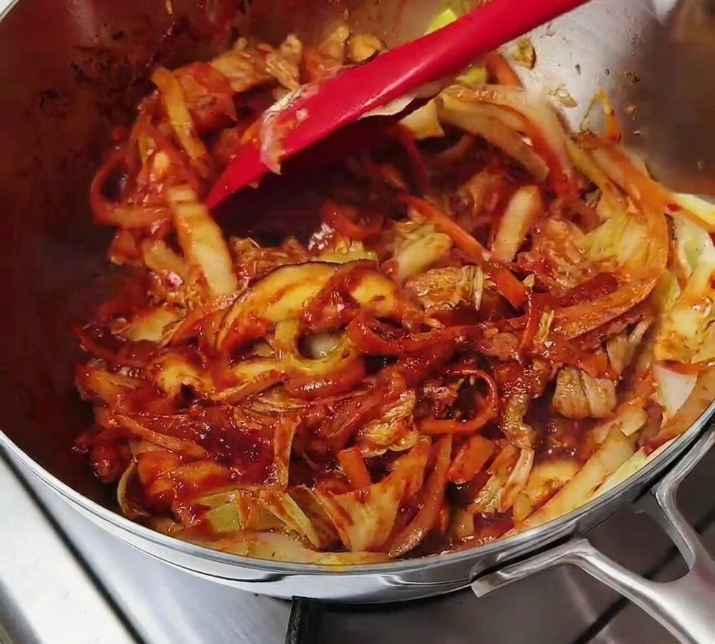 Espátula roja salteando carne y verduras cubiertas completamente por una pasta de chile roja intensa.