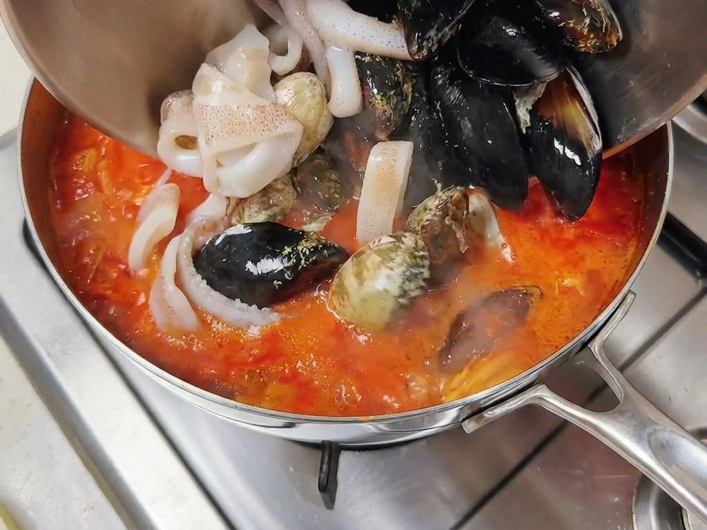Marisco fresco, incluyendo mejillones y anillas de calamar, vertiéndose desde un cuenco al caldo rojo hirviendo.