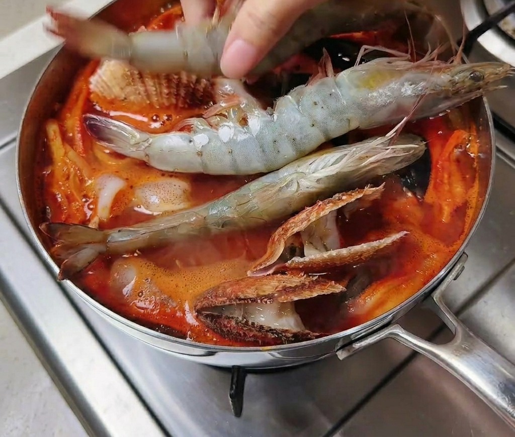 Mano colocando langostinos grandes crudos en una olla de sopa roja hirviendo con marisco.