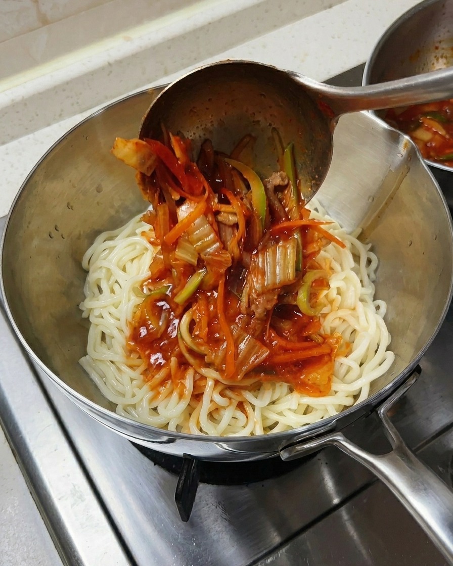 Cazo metálico vertiendo sopa de mariscos y verduras sobre un montón de fideos blancos gruesos en un bol.