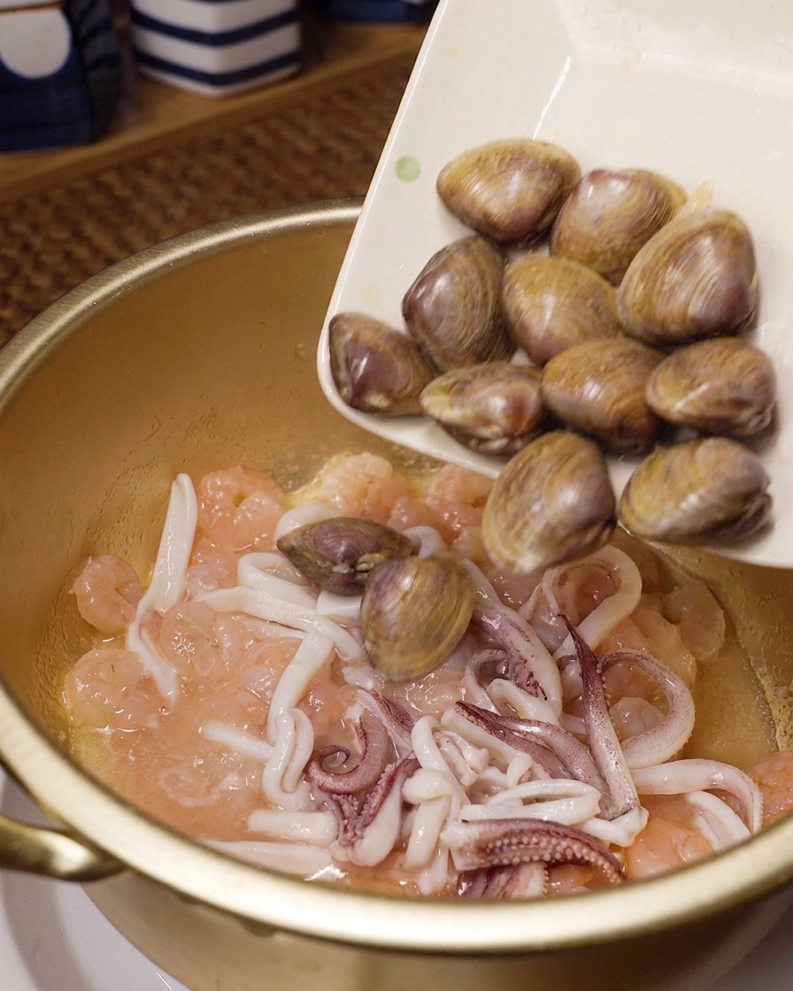Almejas frescas vertiéndose desde un plato blanco en una olla dorada con langostinos y calamares salteados.