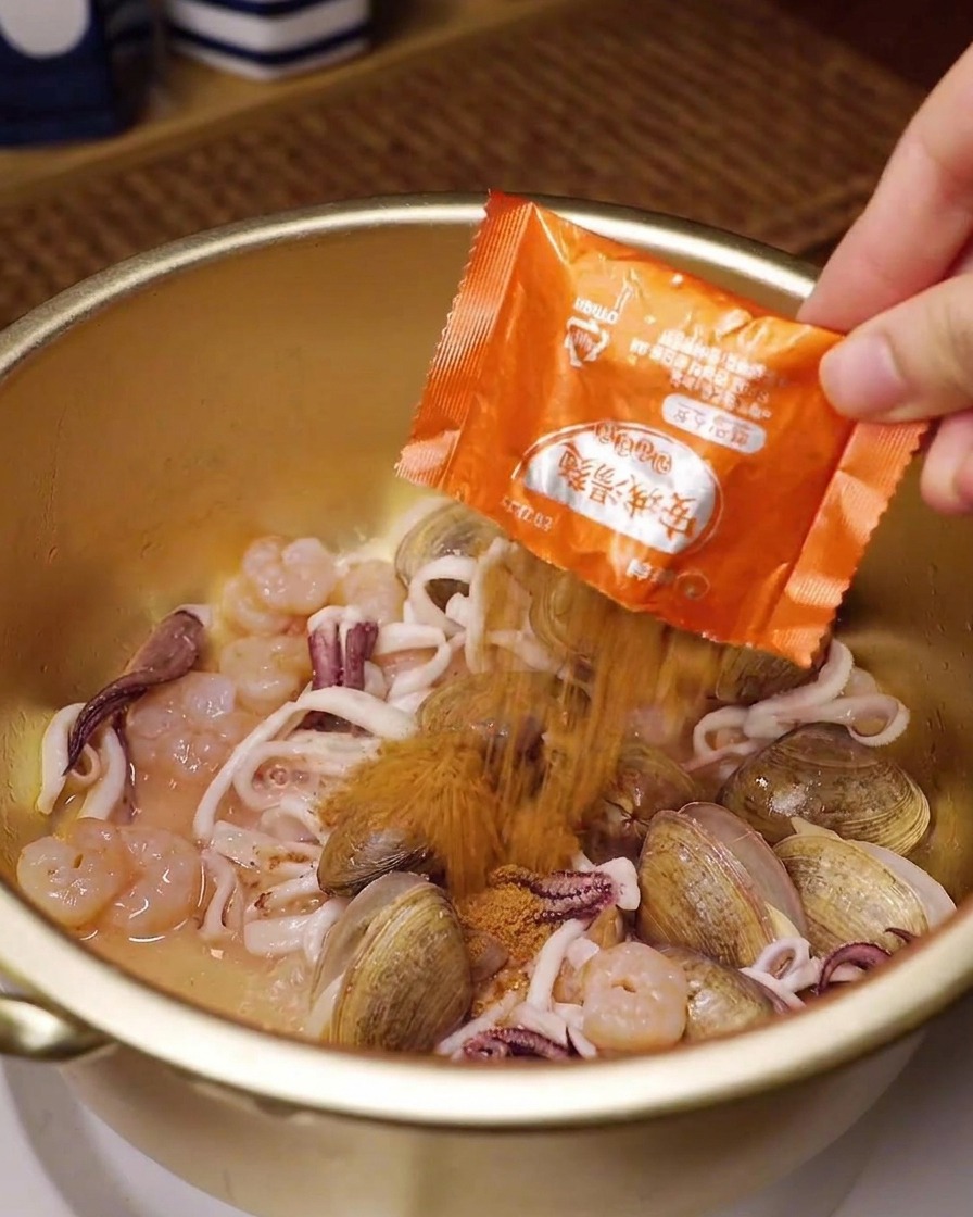 Una mano vertiendo el sobre naranja de condimento para ramen sobre almejas, calamares y langostinos en una olla de latón.
