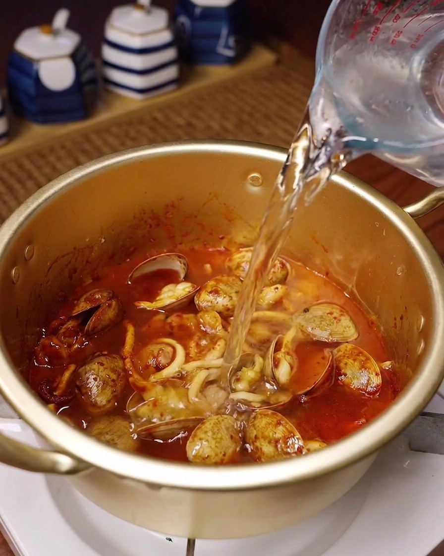 Agua fresca vertiéndose desde una jarra de cristal en una olla dorada con el marisco salteado y especiado.