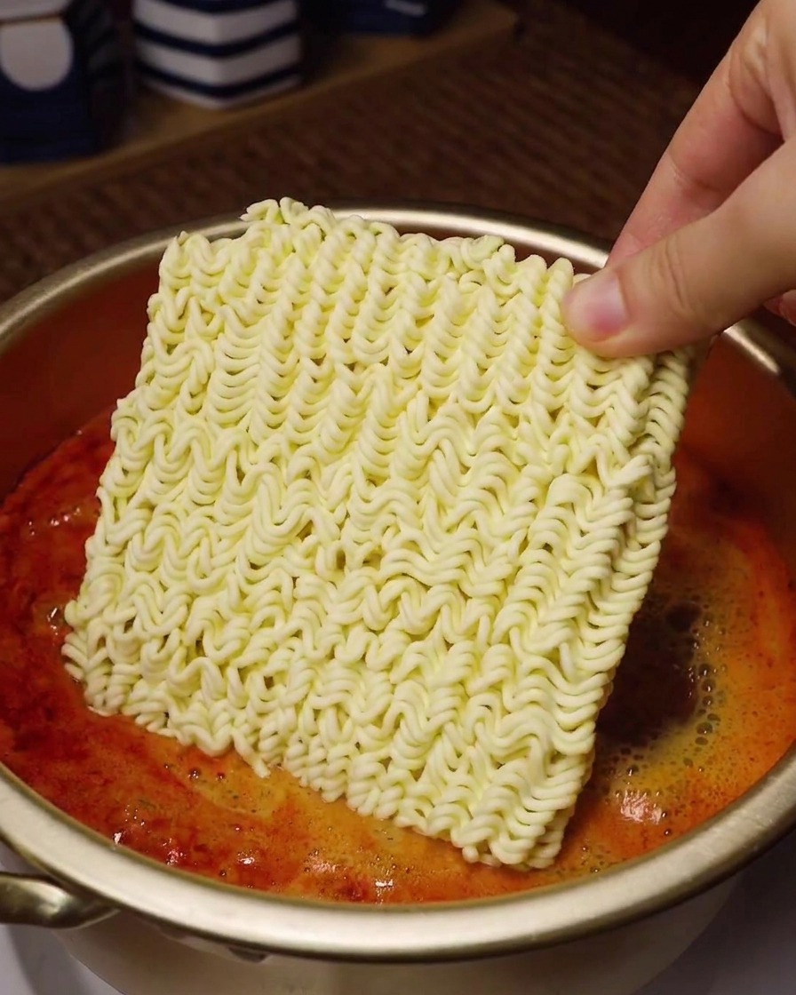 Una mano sosteniendo el bloque de fideos secos sobre una olla dorada con sopa roja hirviendo.
