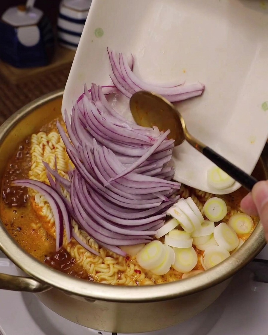Rodajas de cebolla roja y cebolleta vertiéndose en una olla de ramen humeante con caldo rojo.