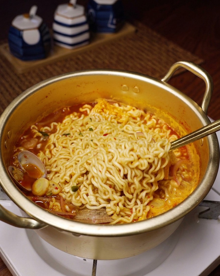 Fideos ondulados levantándose del caldo rojo de mariscos con una cuchara de metal.
