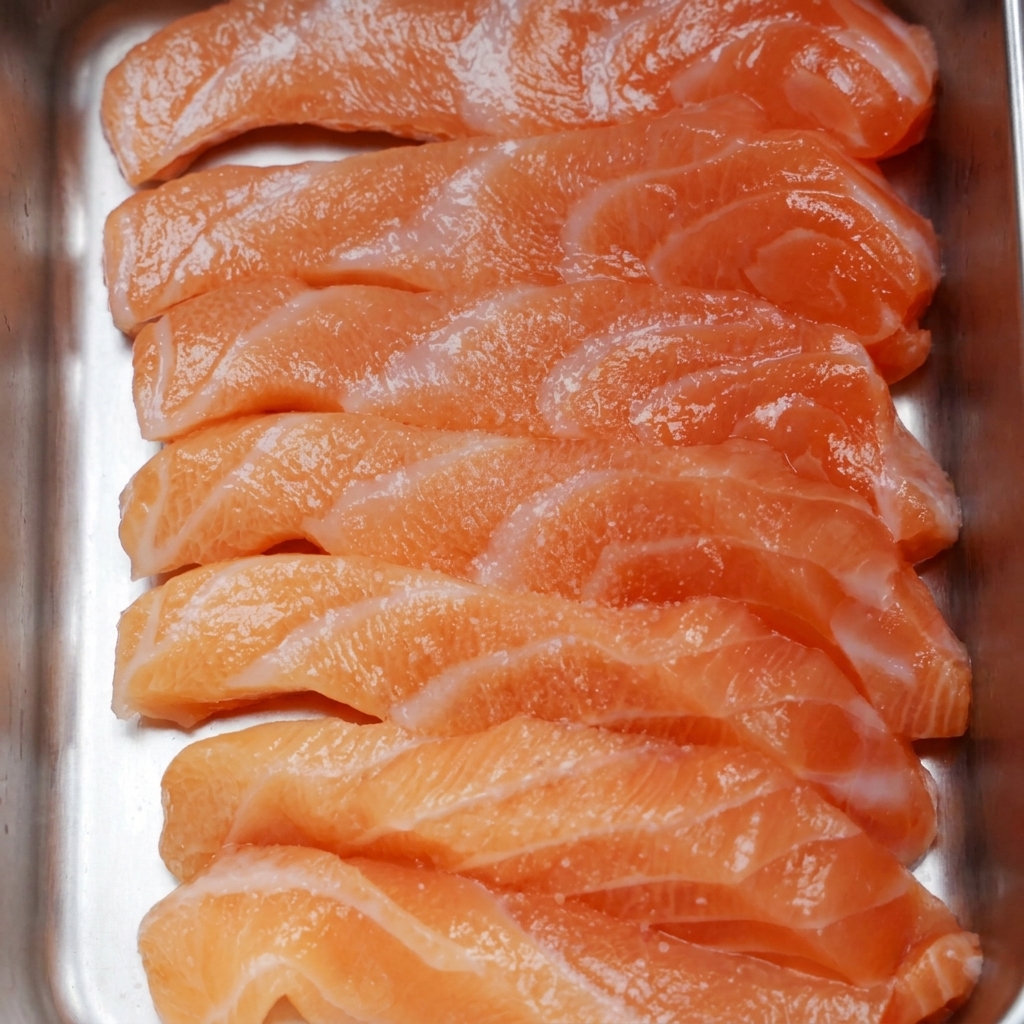 Filetes de salmón crudo en una bandeja metálica, espolvoreados con sal fina y pimienta negra.