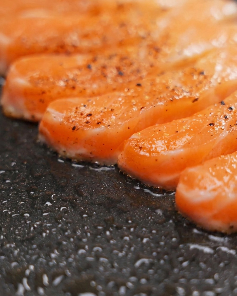 Filetes de salmón sazonados dorándose en una sartén antiadherente con pimienta visible en la superficie.