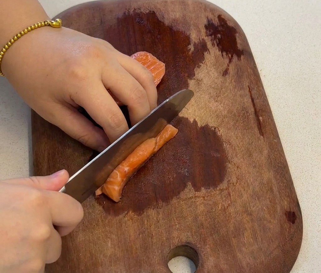 Una mano usando un cuchillo para cortar un filete de salmón fresco en cubos pequeños y uniformes sobre una tabla de madera.