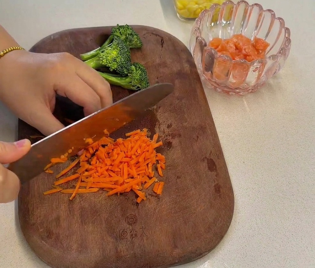 Zanahorias ralladas siendo picadas finamente con un cuchillo de chef sobre una tabla de madera junto a ramilletes de brócoli.