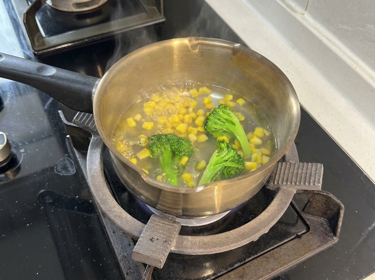 Una olla de acero inoxidable con agua hirviendo, ramilletes de brócoli y granos de maíz amarillos.