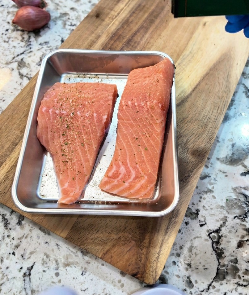 Una mano sazonando filetes de salmón crudos en una bandeja con una pizca de pimienta negra y ajo en polvo.