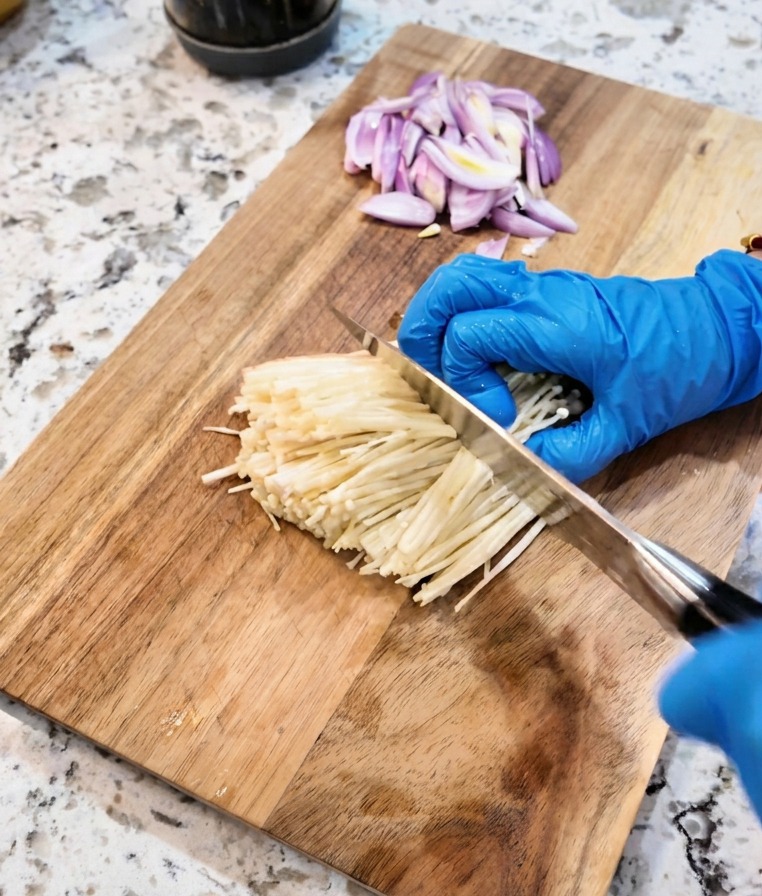 Un cuchillo cortando la base de un manojo de setas enoki blancas sobre una tabla de cortar.