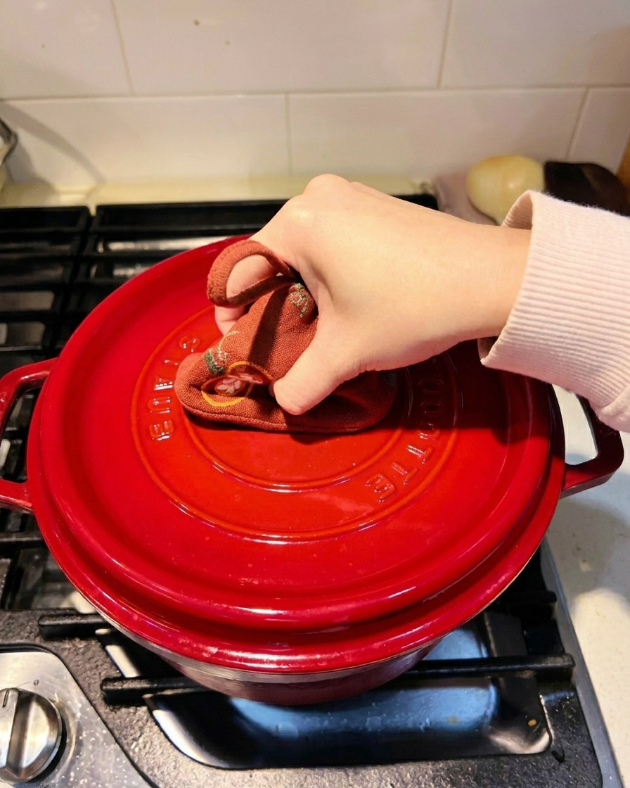 Una mano usando un paño de cocina para presionar la tapa de una olla de hierro fundido roja.