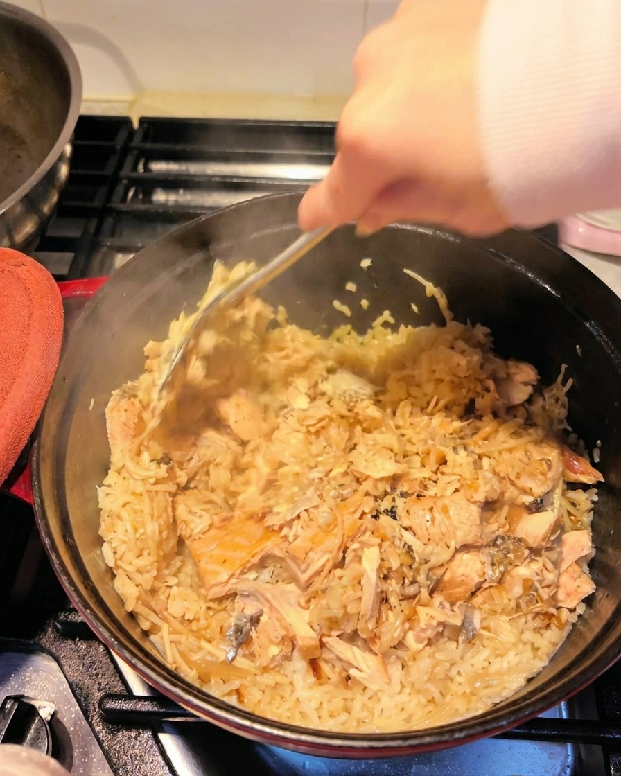 Una mano usando una espátula para trocear los filetes de salmón y mezclarlos con el arroz sazonado.