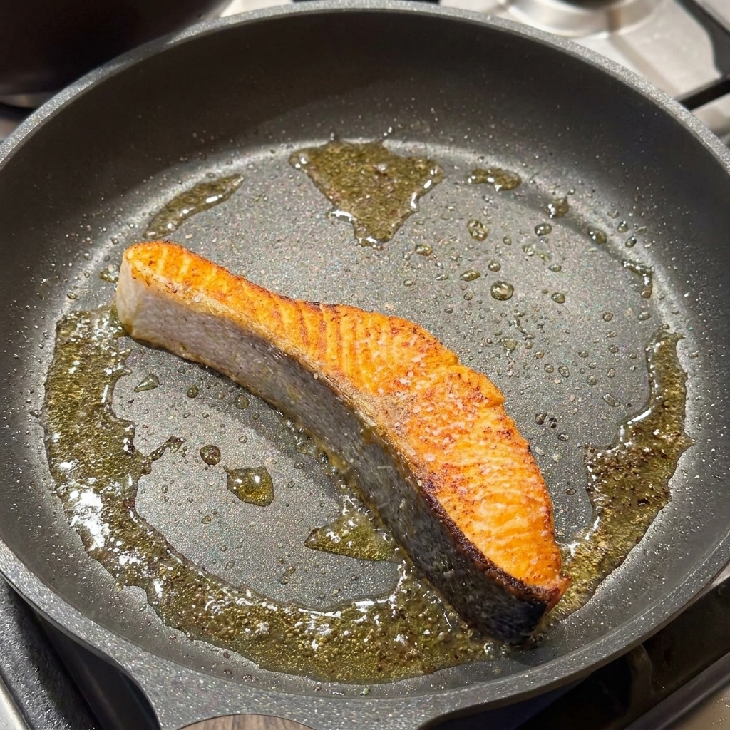 Un filete de salmón sellado en una sartén siendo sazonado con sal y pimienta negra.
